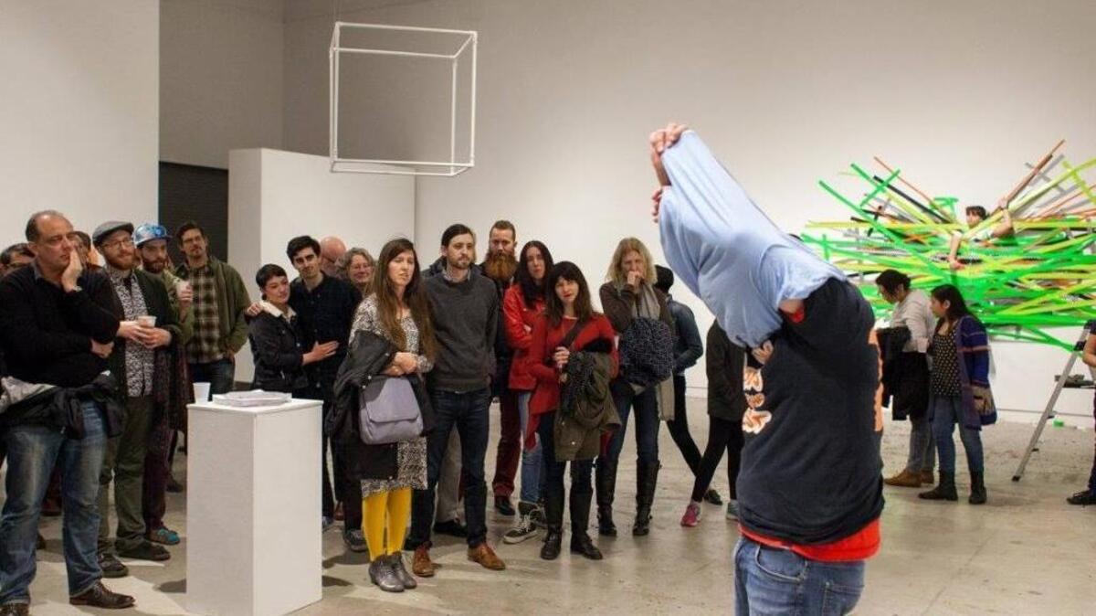 Connor Mickiewicz interprets Jerome Bel’s “Shirtology,” a T-shirt striptease, at the “do it” opening at Verge Center for the Arts. In the background at right is Robert Ashley’s piece involving the duct-taping of a Verge intern several feet up a wall, where she hung for 45 minutes. And in the background at left is Mel Bochner’s “Territorial Reserve #4,” a cube made of PVC pipe and hung from the ceiling.