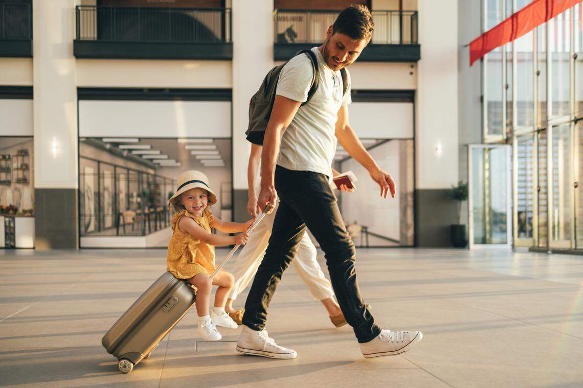 Cheerful husband and his wife walking with their little girl sitting on luggage at the airport.