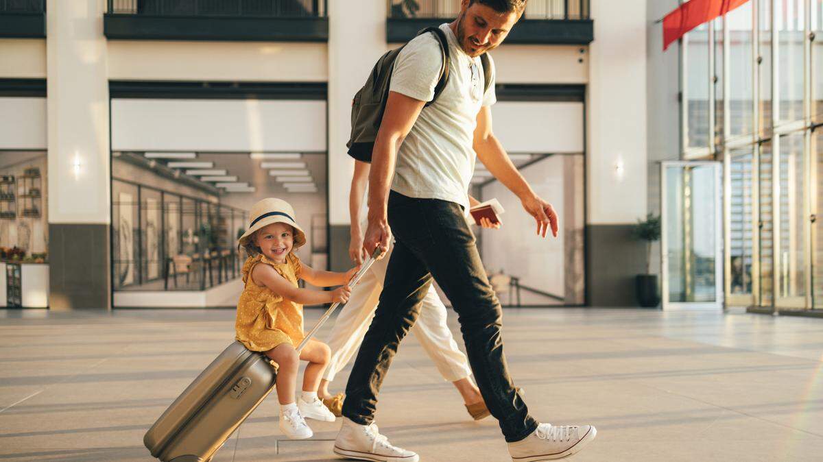 Cheerful husband and his wife walking with their little girl sitting on luggage at the airport.