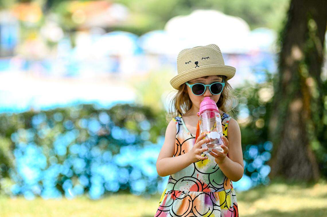 A young girl drinking water out of a reusable bottle with a sun hat and sunglasses on.