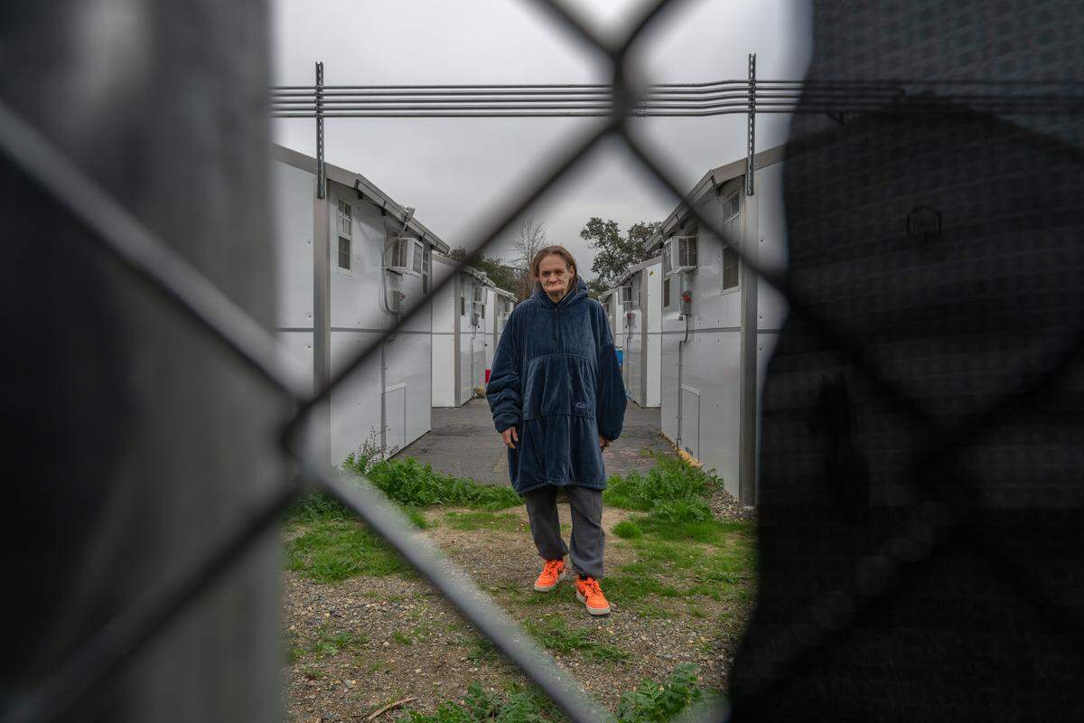 Susan Alhaqq, 54, is photographed through a gap in a fence near a row of sleeping cabins where she now lives with her husband at the Roseville Road shelter in Sacramento earlier this month. She has been living at the shelter for the past year and a half and secured a space with air-conditioning and heat only four months ago. Before that, she lived in a trailer with mold so severe that she said her husband was hospitalized three times with lung infections.