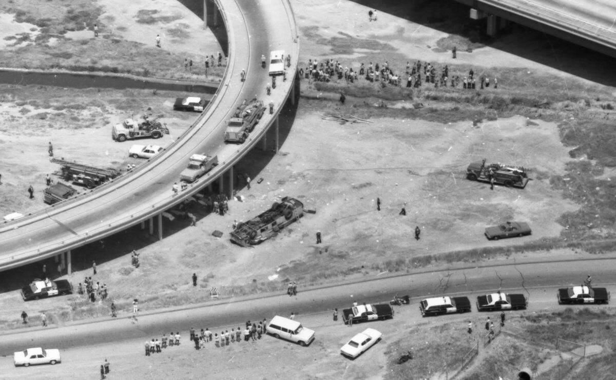 Rescue workers and bystanders surround a Yuba City High School bus that lies on its crushed top below the freeway offramp from which it plunged on May 21, 1976. Bodies of some of the 29 victims are covered by white sheets in the shade beneath the offramp. 