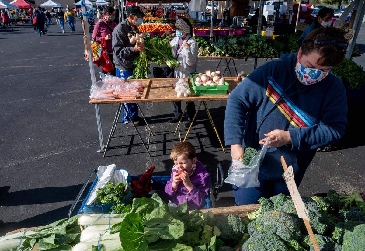 Oliver Schaffer, 4, bites into an apple as his mother Hannah Schaffer, 38, of Sacramento, looks at produce at the Certified Farmers Market in the parking lot of Arden Fair mall in Sacramento on Sunday, March 21, 2021. The market will remain in the mall parking lot until it’s safe to return underneath the W-X freeway.