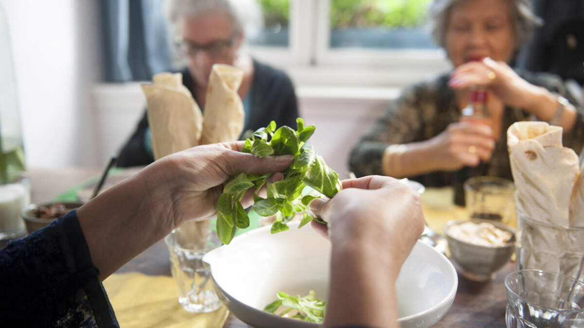 A woman preparing a salad as two other women sit along the other side of the table.