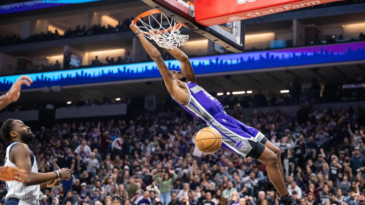 Sacramento Kings guard De’Aaron Fox (5) swings on the rim after a slam dunk past Dallas Mavericks guard/forward Theo Pinson (1), left, during the second quarter at the NBA basketball game Saturday, Feb. 11, 2023, at Golden 1 Center in Sacramento.