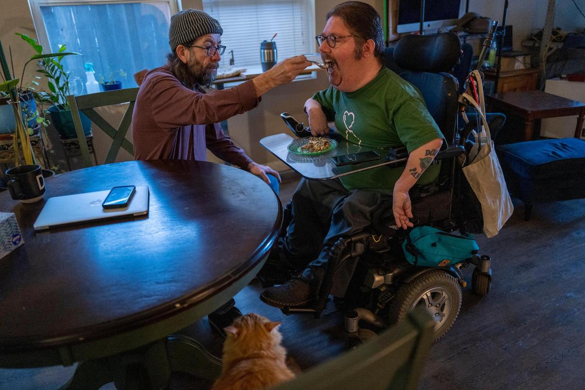 Russell Rawlings, de 46 años, a la derecha, un activista de los derechos de los discapacitados que vive con parálisis cerebral y depende de los cuidadores de Servicios de Apoyo a Domicilio de California, recibe el desayuno de su cuidador Darrow Sprague, de 54 años, el martes. Sprague, quien vive y comparte los gastos del apartamento de alquiler, ayuda a Rawlings en todo, desde la preparación de las comidas y las tareas domésticas hasta la higiene personal, así como a subir y bajar de la cama con un elevador .