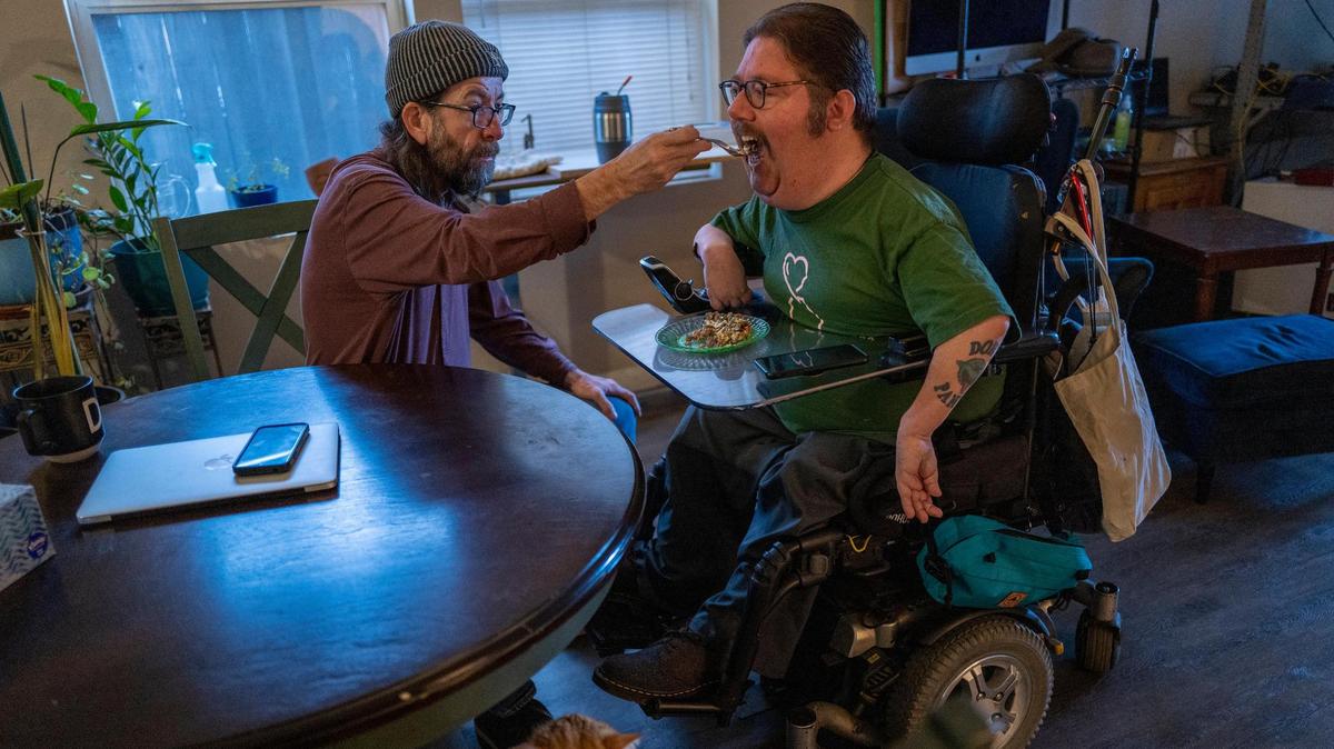 Russell Rawlings, 46, right, a disability rights advocate who lives with cerebral palsy and relies on caregivers from California’s In-Home Supportive Services, is fed breakfast by his caregiver Darrow Sprague, 54, on Tuesday, Feb. 14, 2023. Sprague, who lives in and shares the expense of the rental apartment, helps Rawlings with everything from meal preparation and housekeeping to personal hygiene, as well as getting in and out of bed with a lift.