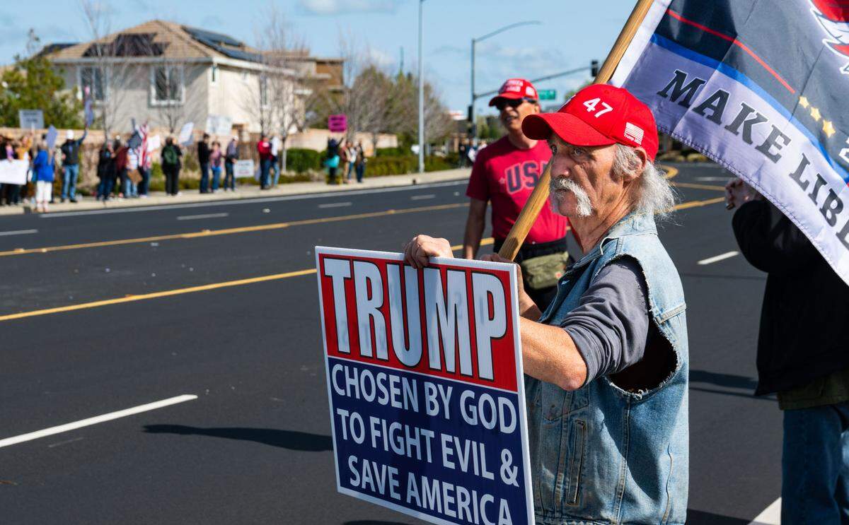 A man with sign that says President Trump was “chosen by God to fight evil & protect America” stands in front of the Tesla dealership as anti-Tesla protesters demonstrate across the street Saturday, March 29, 2025.