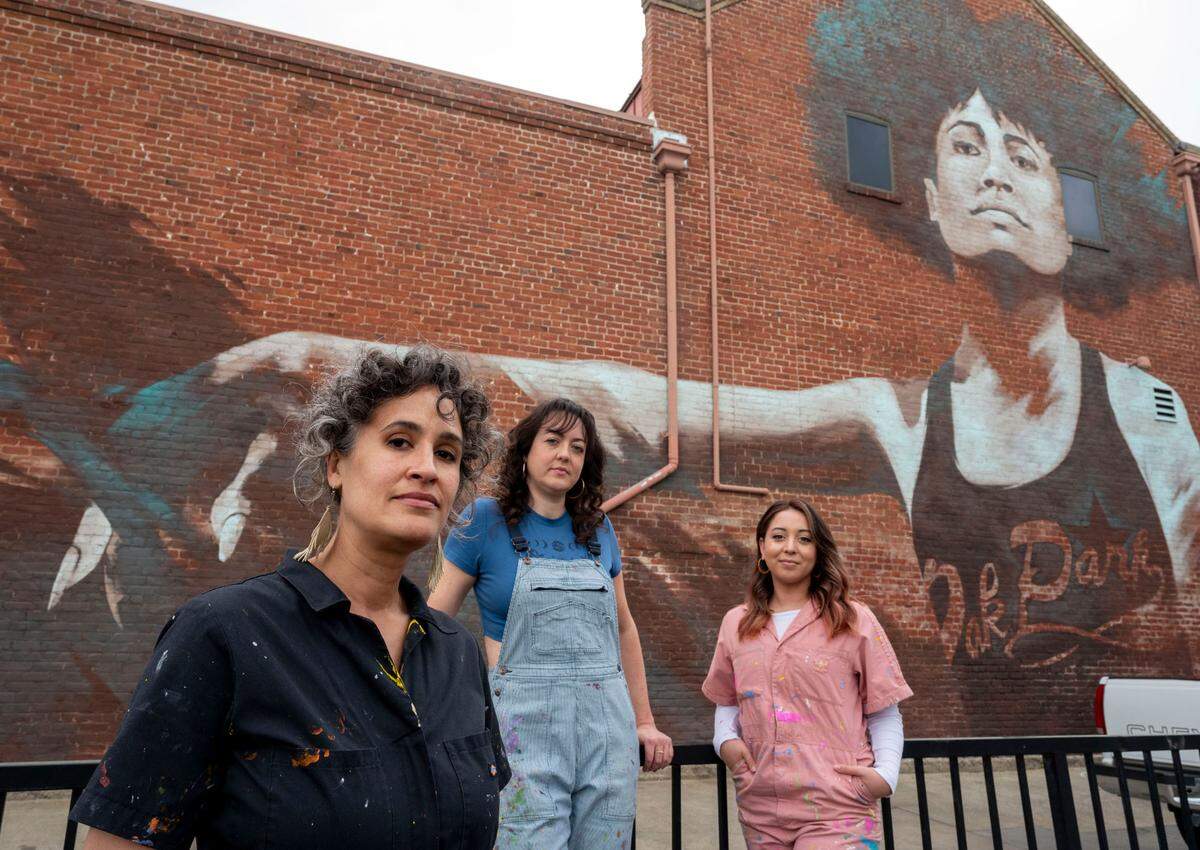 Sacramento artists, Jaya King, left, Jolene Rose Russell, center, and Uli Smith stand in front of a mural March 22 titled “Guild Goddess,” painted by King in 2018 on Oak Park’s Guild Theater for Wide Open Walls. They claim Wide Open Walls exploits new artists by underpaying them.
