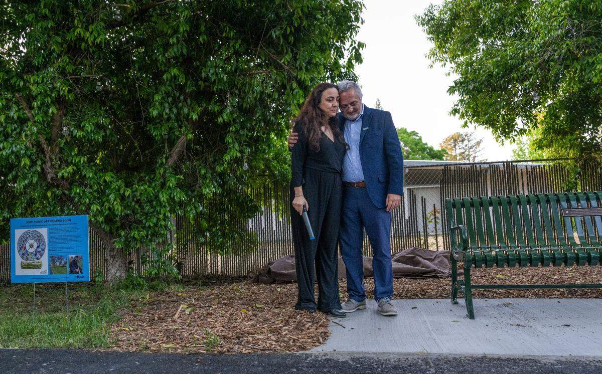 Nadine Yehya, left, and father Majdi Abou Najm stand on Monday near a bench at Sycamore Park in Davis that memorializes their son Karim Abou Najm, a UC Davis student who was fatally stabbed one year ago, during a “day of remembrance” of the event. At left, a sign depicts a sculpture that will be installed at the location in his memory.