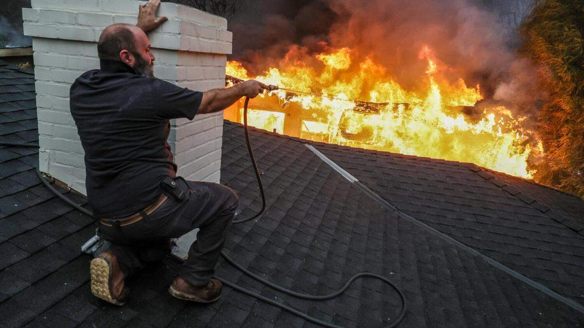 Jimmy Orlandini shields himself from intense heat as he hoses down a neighbor’s rooftop on Sinaloa Avenue in Altadena as the Eaton Fire burned Wednesday.