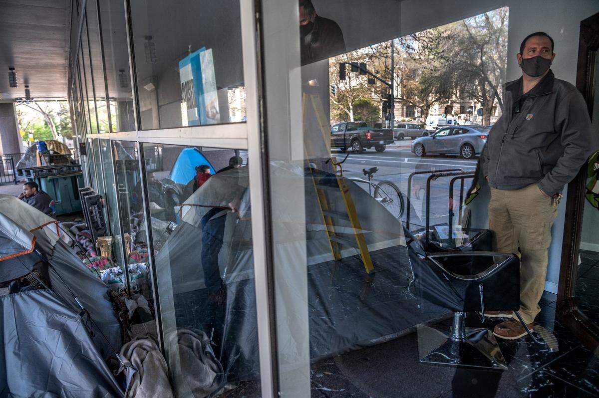 Workers inspect the fire damage inside of Michael Z Salon, owned by Alma Perez, as a homeless encampment is reflected in the window, Wednesday, March 24, 2021. The fire was started in a van above in the City Hall Garage where homeless living in cars were permitted overnight parking.