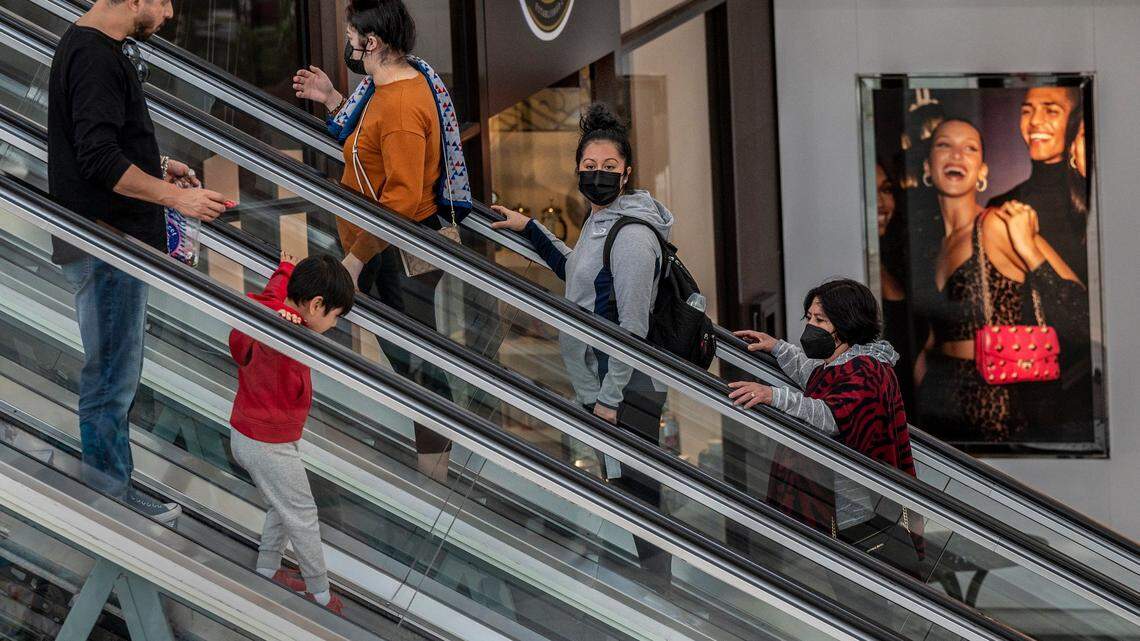 Shoppers, some with masks and some without, ride the escalator at Sacramento’s Arden Fair mall near an advertisement of smiling models outside the Michael Kors store on Tuesday. California’s indoor mask mandate ends Wednesday, but teachers and students must keep masking up according to state guidelines.