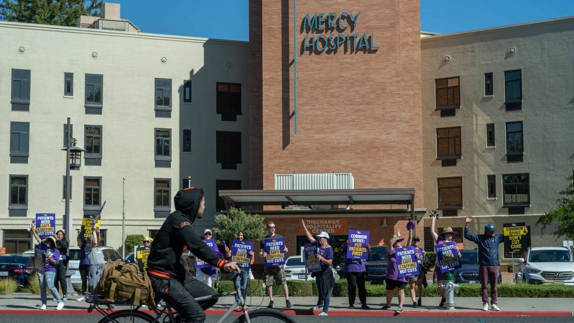 A cyclist watches as CommonSpirit healthcare workers picket outside Dignity Health Mercy General Hospital in Sacramento on Thursday, Sept. 26, 2024, in response to the outsourcing of critical jobs, layoffs and the impact on patient care. California Nurses Association announced Friday that Dignity Health nurses have ratified a new four-year contract.
