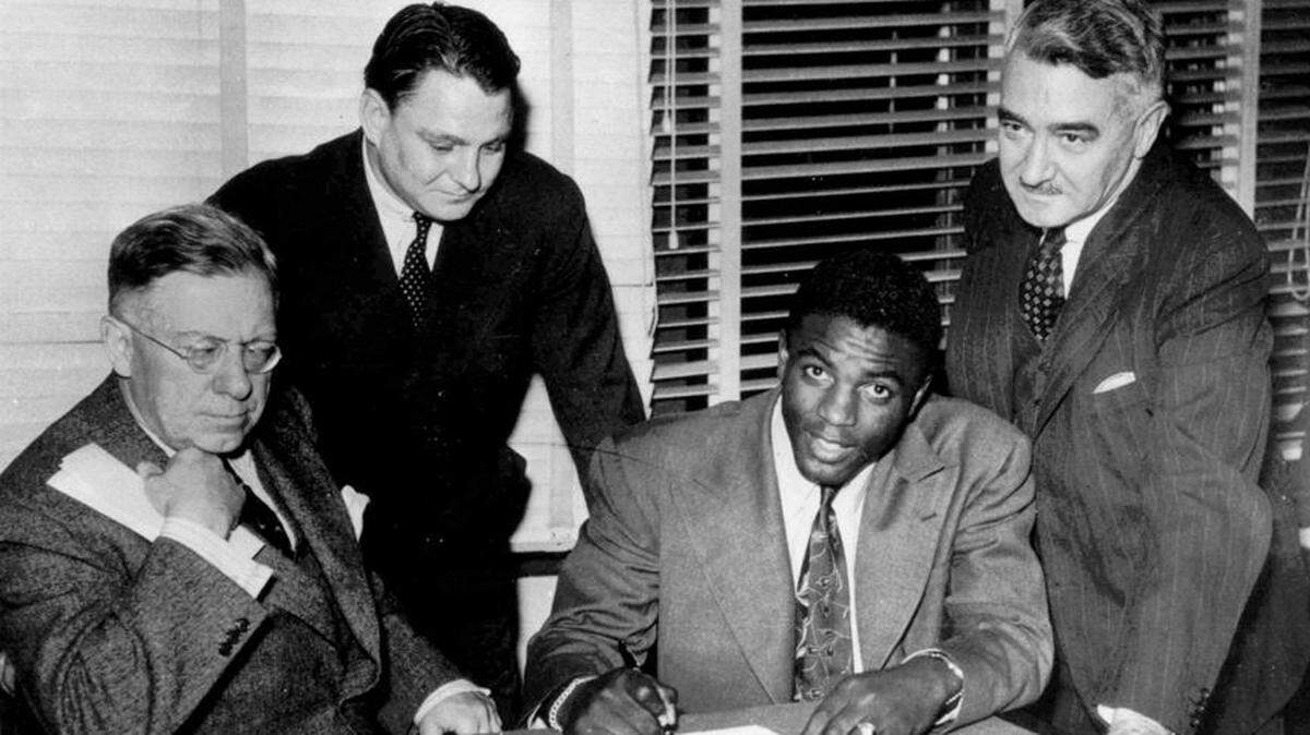 Dodgers staffer Branch Rickey Jr., left, watches as Jackie Robinson signs his historic contract on April 11, 1947.