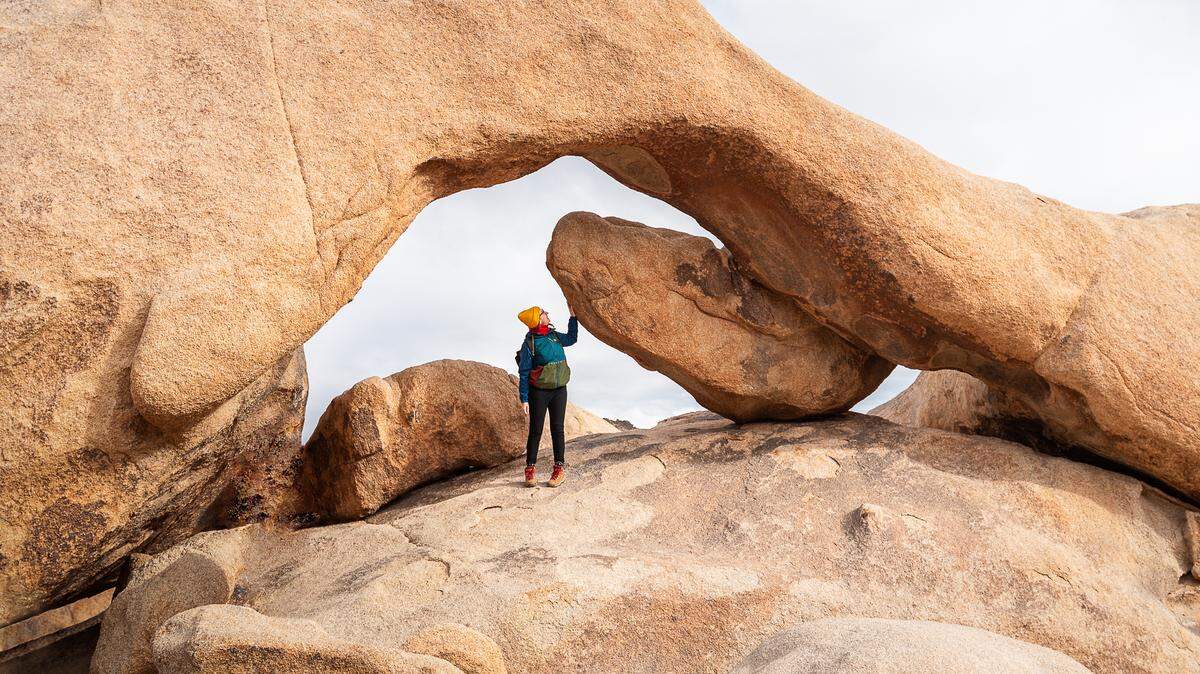 Woman standing under a huge yellow-orange rock formation, which looks like a tunnel. Only the sky is visible in the background. The location is Joshua Tree National Park in Southeastern California.