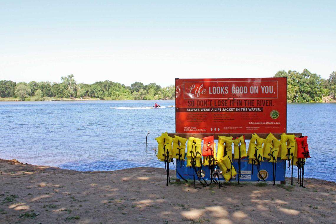 Firefighter Ross Palmerton of the Sacramento Fire Department assists in a mock rescue behind a life jacket rack and sign on Tiscornia Beach, part of the American River, on Tuesday, May 23, 2023. Ahead of Memorial Day weekend, city officials from the office of emergency management, fire department and youth, parks and community enrichment hosted demonstrations including drowning rescue and CPR administration.