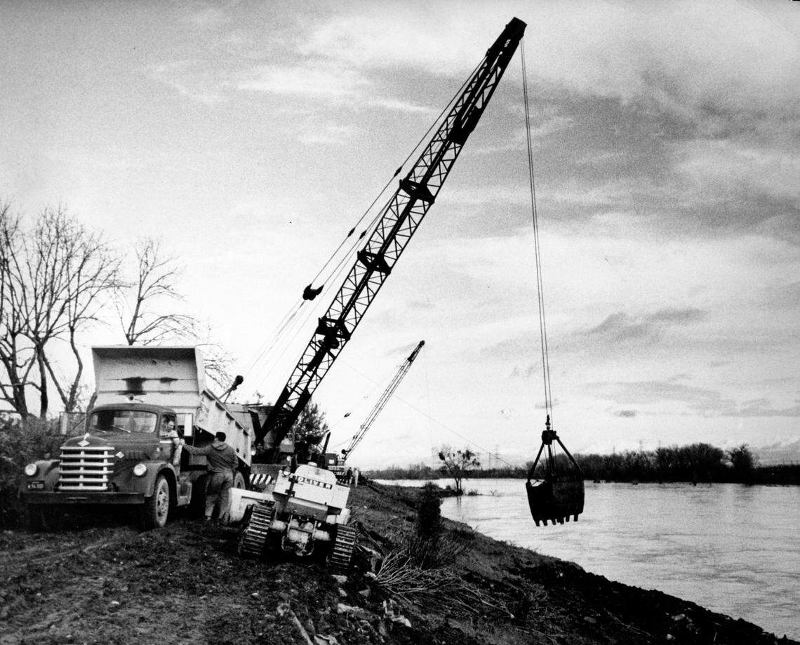 A clam shell crane places 60 tons of rock and earth fill on the River Park levee, downstream from Sacramento’s H Street Bridge, in December 1964, as the American River runs past at a high level. The fill, which included concrete from the old Edmonds Field baseball grandstand, was meant to address observed erosion on the levee bank.