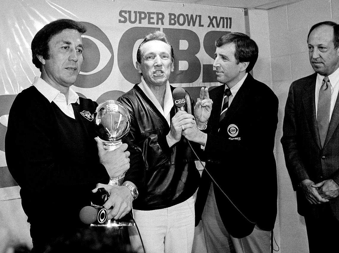 In this Jan. 23, 1984, photo, Oakland Raiders coach Tom Flores clutches the Super Bowl trophy as managing general partner Al Davis is interviewed by Brent Musburger in the locker room after a 38-9 win over the Washington Redskins in Super Bowl XVIII in Tampa, Fla. At right is NFL Commissioner Pete Rozelle.