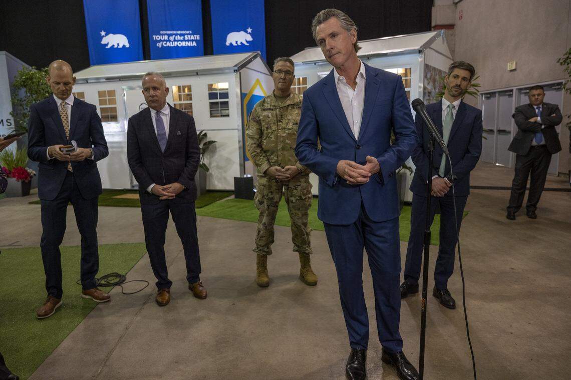 After touring tiny homes at Cal Expo, Gov. Gavin Newsom answers questions from the media on March 16, 2023, after announcing that the state will have 1,200 of the homes built and delivered throughout California to help house the homeless population. Kevin McCarty, then a member of the Assembly, stands at left with then-Sacramento Mayor Darrell Steinberg and California National Guard Maj. Gen. Matt Beevers, center.