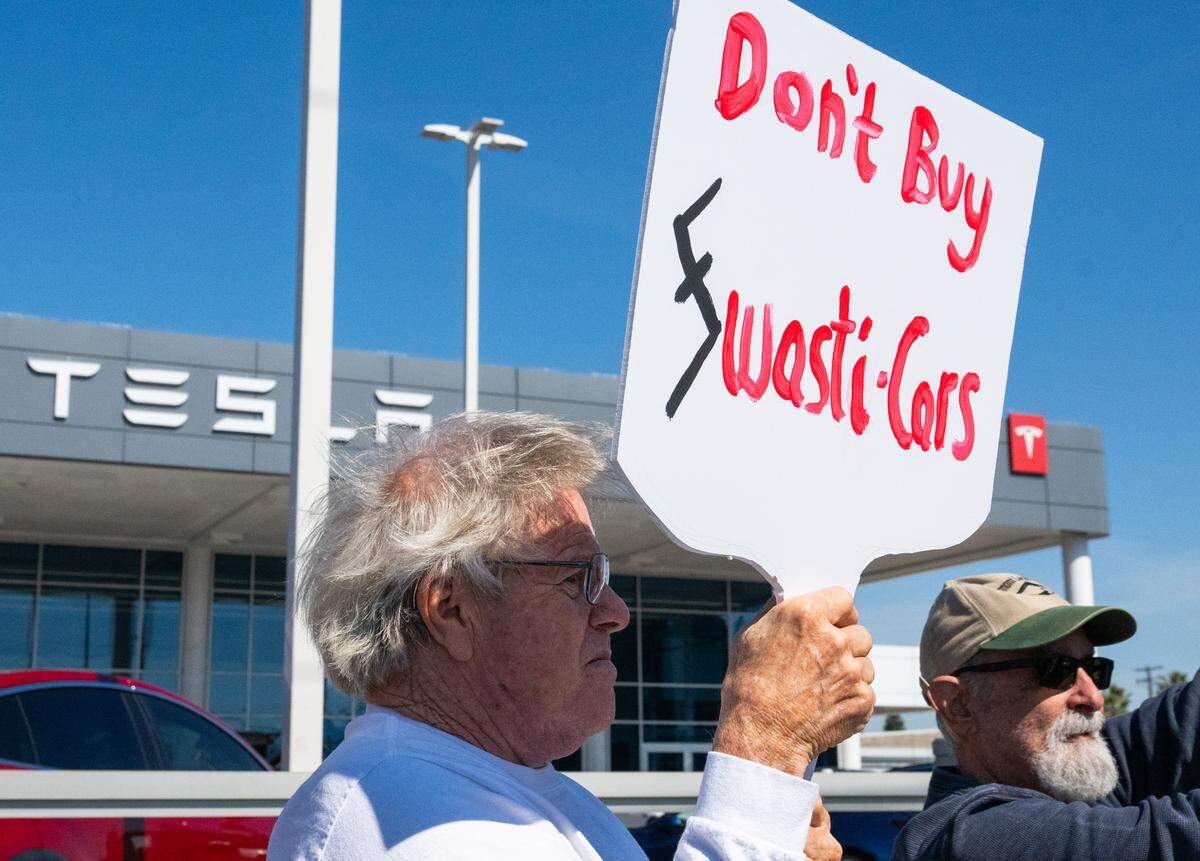 “Don’t buy swasti-cars” reads a sign held by Steve Davenport during a protest against Elon Musk at a Tesla dealership on Arden Way near Sacramento on Saturday, March 8, 2025. More than 100 people lined both sides of street to protest the role of the tech billionaire and Tesla CEO in the Trump administration.