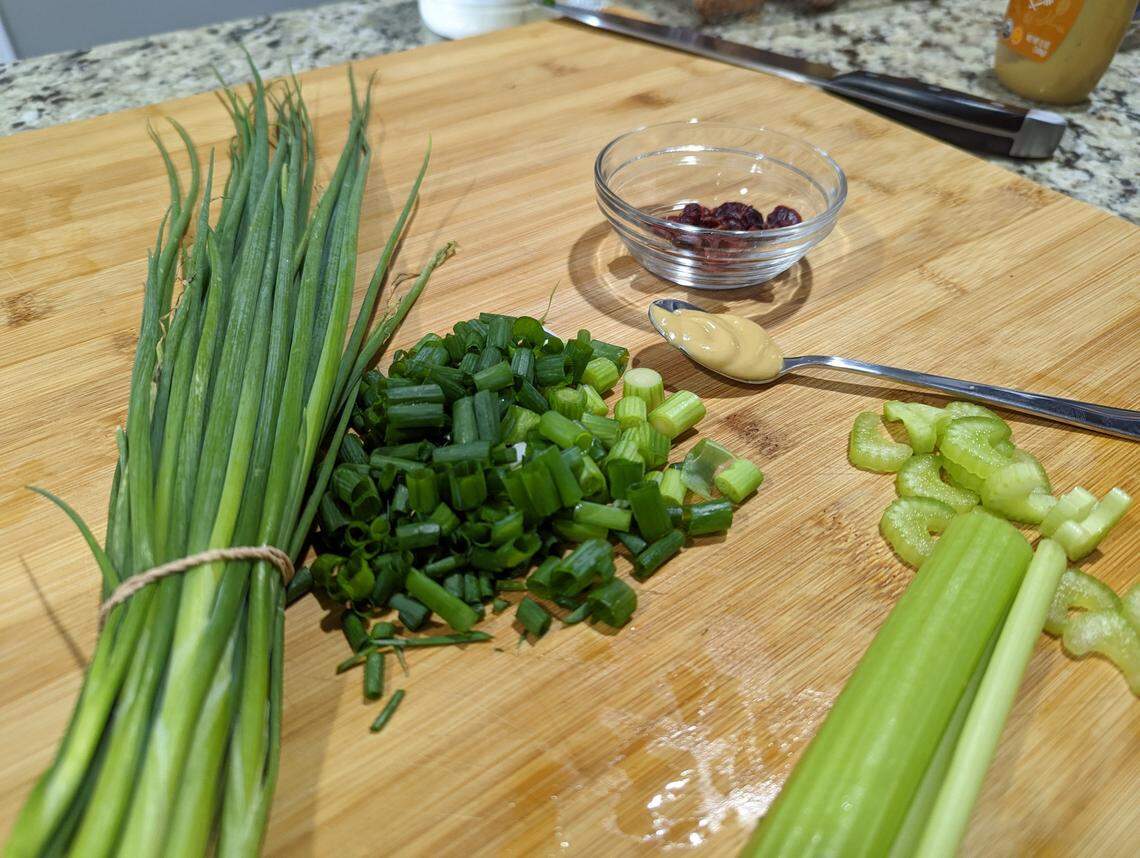 Green onions and celery on a cutting board, along with a spoonful of honey mustard and a small bowl of dried cranderries.