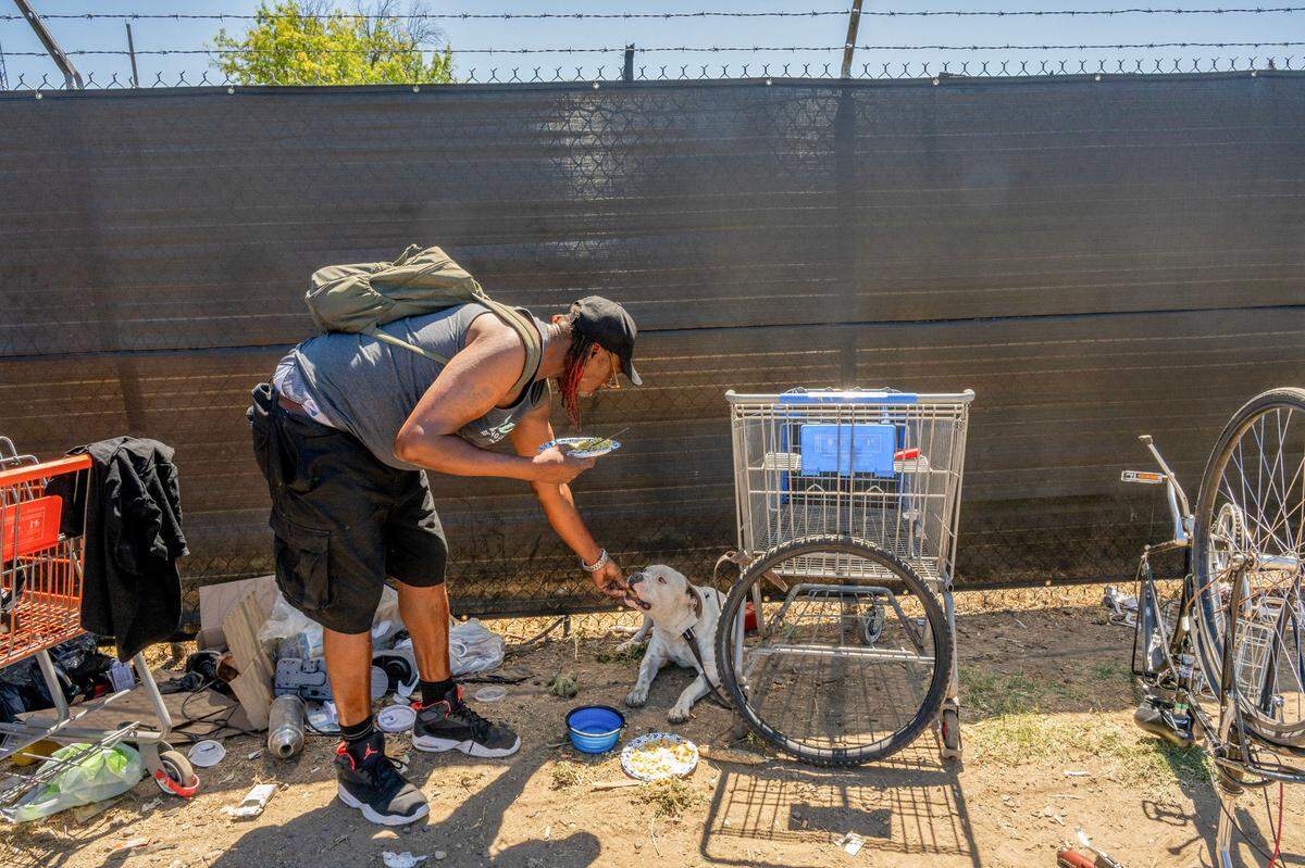 George Robinson shares his lunch with his dog Kane outside the fence of First Step Communities Campus for the homeless where he lives in a pallet home on Roseville Road in Sacramento on Wednesday, Aug. 21, 2024.