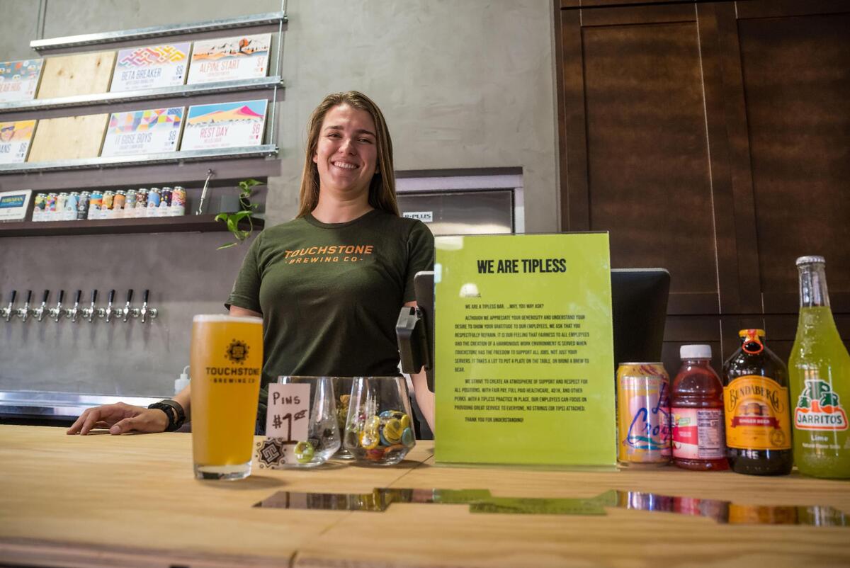 Kristen Madigan, pub manager at Touchstone Brewing Company, stands behind the bar Friday, Aug. 18, 2023, in Sacramento. A sign next to the payment area explains why their establishment is “tipless.”