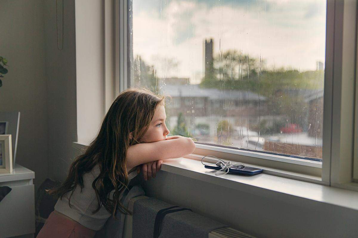 A girl sits alone at the window of an apartment. What is the age requirement for children to be home by themselves in California? How old? How young is too young?