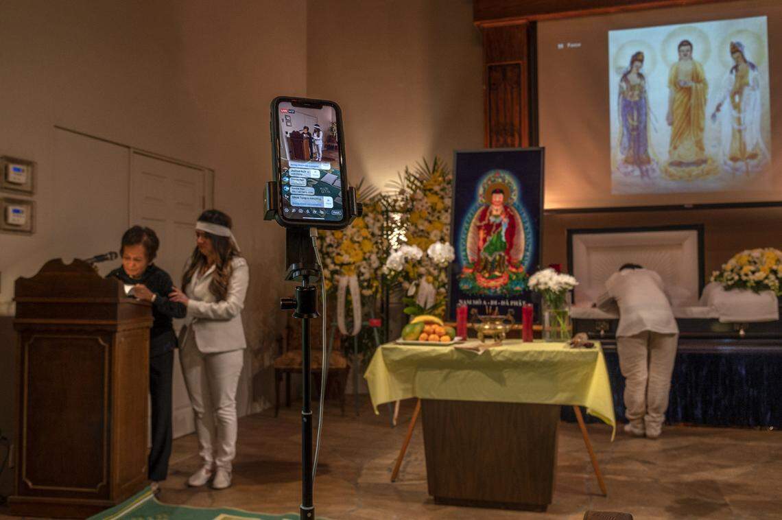 A live stream recording is set up so family and friends of Dung Tan Nguyen can view his funeral service from around the world, Wednesday, May 6, 2020, as family members abide by restrictions put in place to prevent the spread of the coronavirus. At left, Trang Nguyen supports her husband’s aunt Kim Anh Dinh as she reads farewell messages to an empty room. At right, son Bao Nguyen leans over his casket during the service. The family said they were torn between grieving and trying to live stream the funeral since only four of them were allowed in the room.