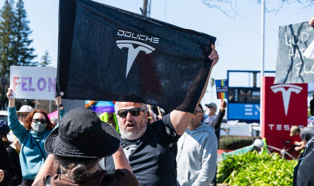 A protestor photographs Ray Shomon and his anti-Tesla flag at a rally against Elon Musk at a Tesla dealership on Arden Way near Sacramento on Saturday, March 8, 2025. More than 100 people lined both sides of street to protest the role of the tech billionaire and Tesla CEO in the Trump administration.