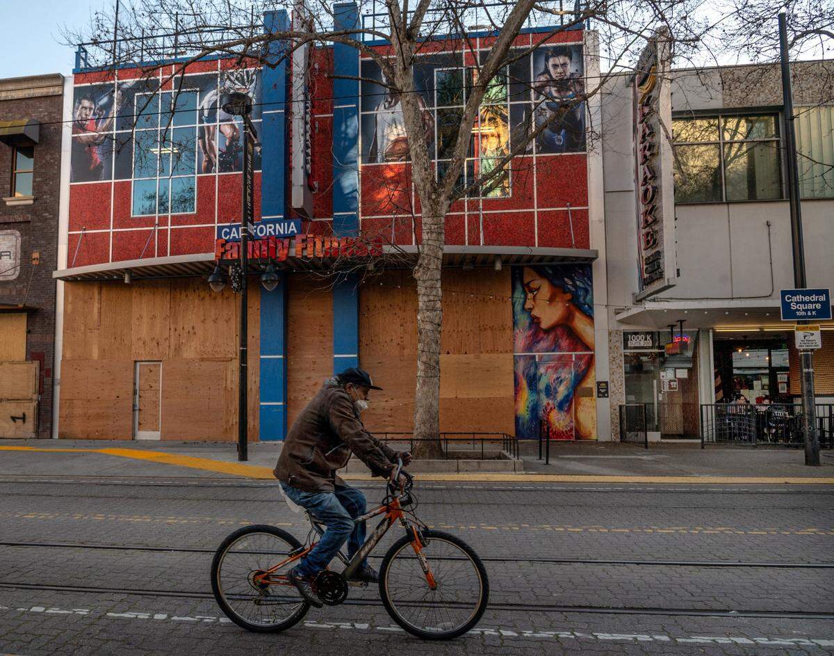 A masked bicyclist rides past the boarded up California Family Fitness, its facade decorated by a Wide Open Walls mural by Charmaine Olivia, on K street in downtown Sacramento in February 2021. Olivia’s mural was later removed.