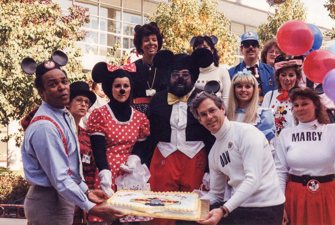Marcy Jacobs, far right corner, celebrates Mickey Mouse’s birthday in November 1988 with coworkers at the California Department of Justice. Don Peri, who is wearing a white turtleneck and helping hold the cake, keeps the photo in his Davis home.