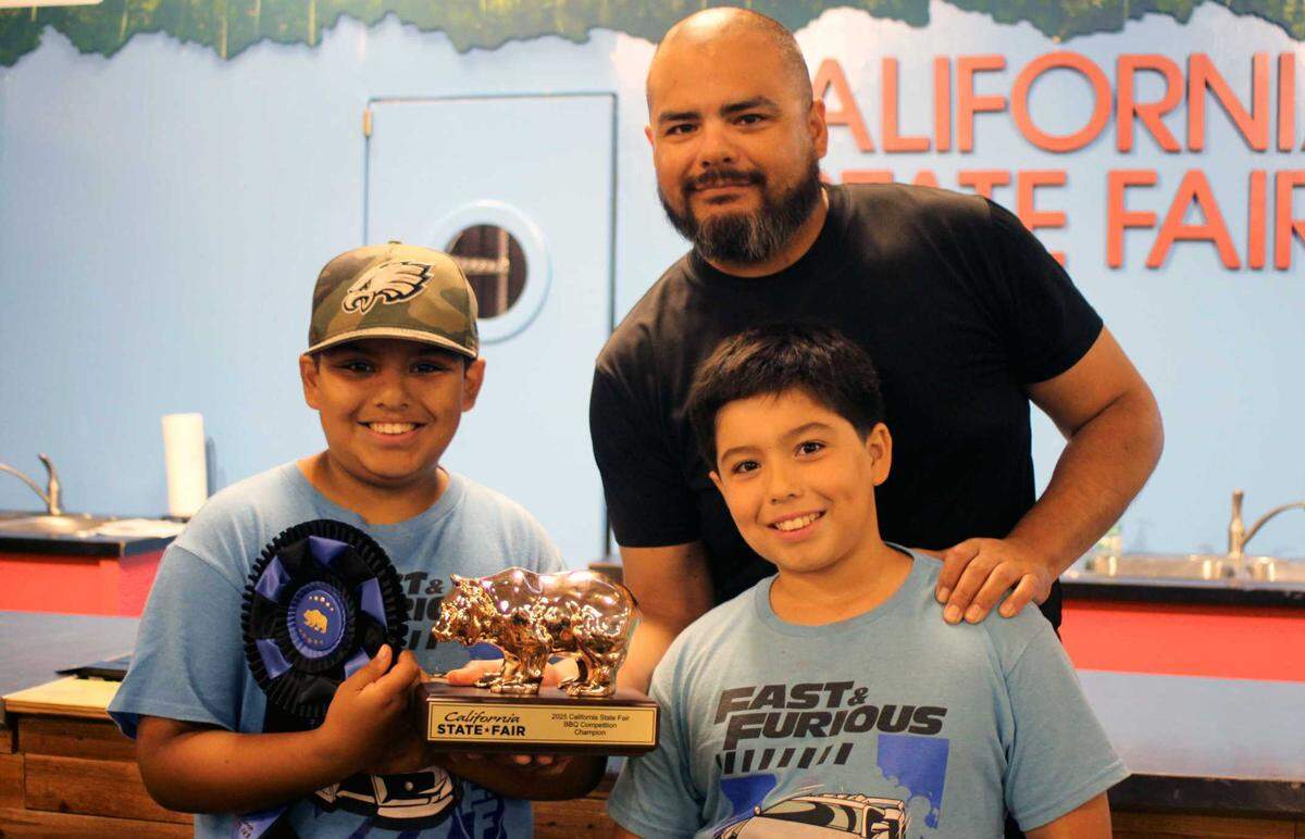 Coaches Pit BBQ pitmaster Jorge Lara (center) wins first place in the California State Fair BBQ Competition and poses with the Golden Bear trophy and his sons Matias Lara (left) and Issac Lara (right) at Cal Expo on July 12, 2025.