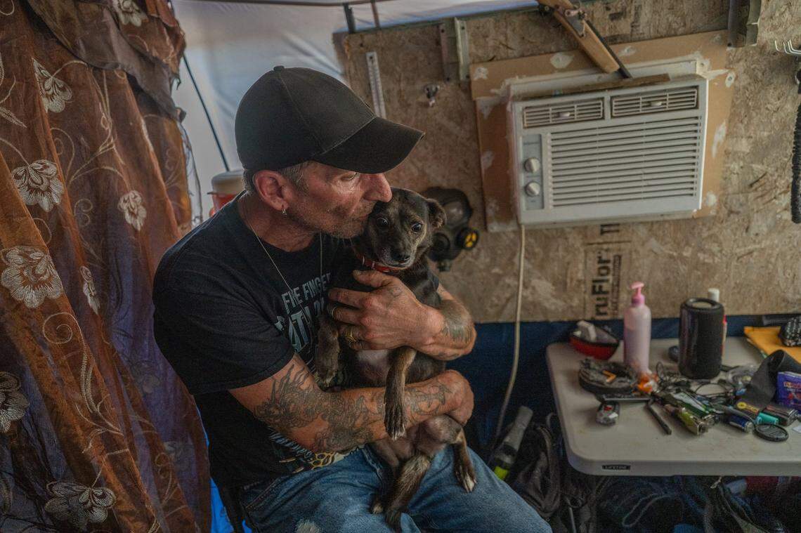 Nikki Buckles, a former carpenter, sits with his dog Opium on Tuesday near an air conditioner he hooked up to a generator to stay cool in a tent and wooden makeshift shelter. He was worried about leaving his possessions to go to a cooling center but said he might go if he felt the dog is in danger from the heat.