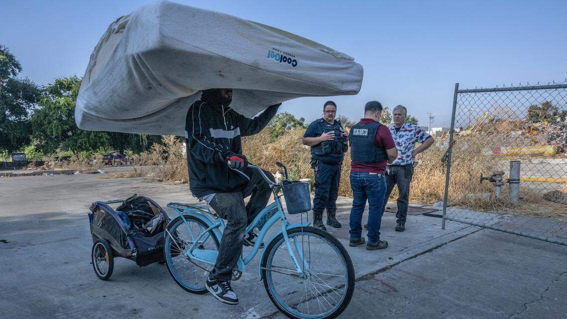 A homeless man carries a mattress as he cycles past city enforcement officers during a homeless encampment sweep on 1st Avenue in Sacramento on Wednesday Sept. 20, 2023. The rubble of the California Shellfish Company building that burned down on Aug. 24 rests at right.