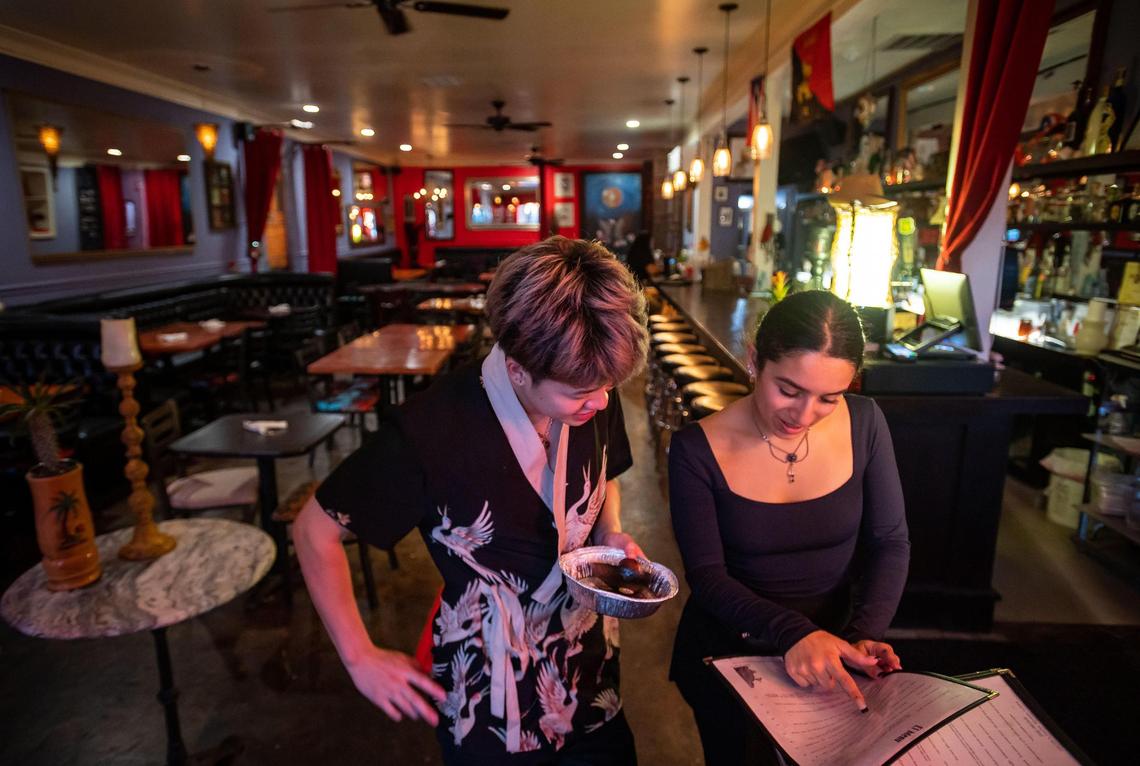 Gerry Hu, left, a sushi chef at nearby Miso Japanese Restaurant, looks at the Lola’s Lounge menu with help from Lola Serrano, who manages and co-owns the restaurant with her mother Annette, in front of an empty dining room last month. Employees in the area often support each other by ordering and sometimes exchanging food with other nearby restaurants around the Broadway district, where business has slowed or shuttered amid concerns surrounding safety.