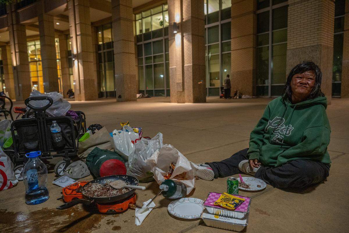 Mane “Jessica” Davila eats dinner she cooked for her diabetic husband, Filipe, as he secures a spot for them to sleep near Sacramento City Hall on Tuesday, Aug. 19, 2025, in Sacramento, just days before an ordinance prohibiting unhoused individuals from sleeping outside it took effect. “My husband is diabetic. I have to make sure he eats healthy — not a lot of junk food. I have to cook fresh meals for him, not canned food all the time,” she said.