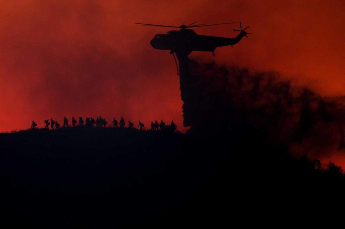 A helicopter drops water as firefighters walk along a ridge during the Sandy Fire in Runkle Canyon on Tuesday, May 19, 2026, in Simi Valley. Cal Fire leaders say their agency has expanded to more than 12,000 firefighters and maintains strong operational coordination with federal crews despite tensions over recent Forest Service staffing cuts.
