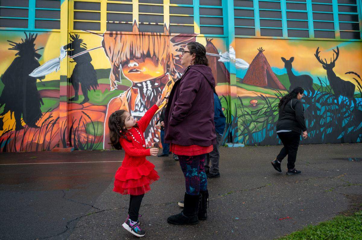 Iris Williams, 4, reaches for her mother Viola Williams, a tribal citizen of the Wilton Rancheria, during the unveiling of a mural on the outside of the gym at Miwok Middle School on Saturday that celebrates its name change from Sutter Middle School.