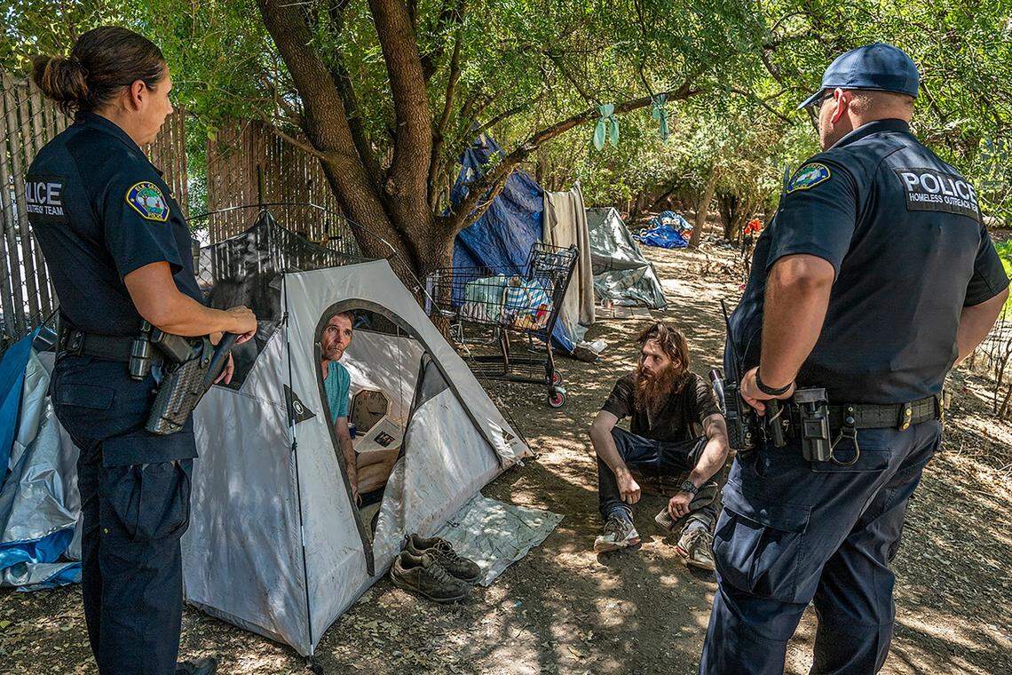 Homeless outreach officers Jennifer McCue, left, and Chris Cahill with the Elk Grove Police Department talk with homeless campers Jason Anduss, inside tent, and Allen Buchman, seated, in Elk Grove on July 19. A new city ordinance will clamp down on encampments. It bars the unhoused from camping within 500 feet of daycare centers, schools, playgrounds or youth centers; bans encampments without permitted electric, water and sewage hookups; and prohibits camping on private property without the property owner’s permission.