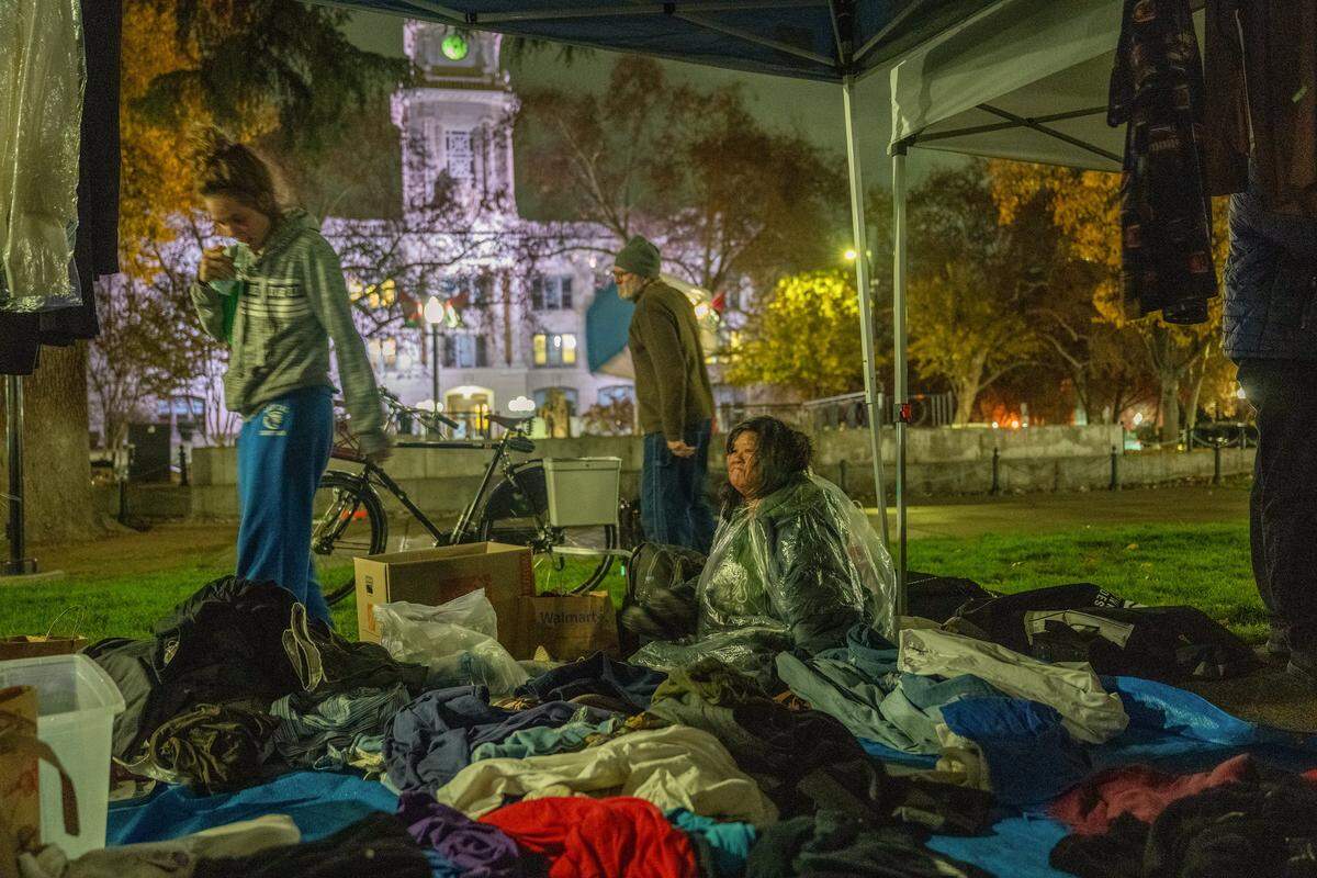 Mane “Jessica” Davila, 56, sits with a rain cover among clothing donated for the homeless at a vigil at Cesar E. Chavez Plaza in Sacramento on Sunday to memorialize people who died this year while unhoused in Sacramento County. Davila said she and her husband have been homeless since July and, although they are staying in a hotel for a few days, wanted to join the community for the event.