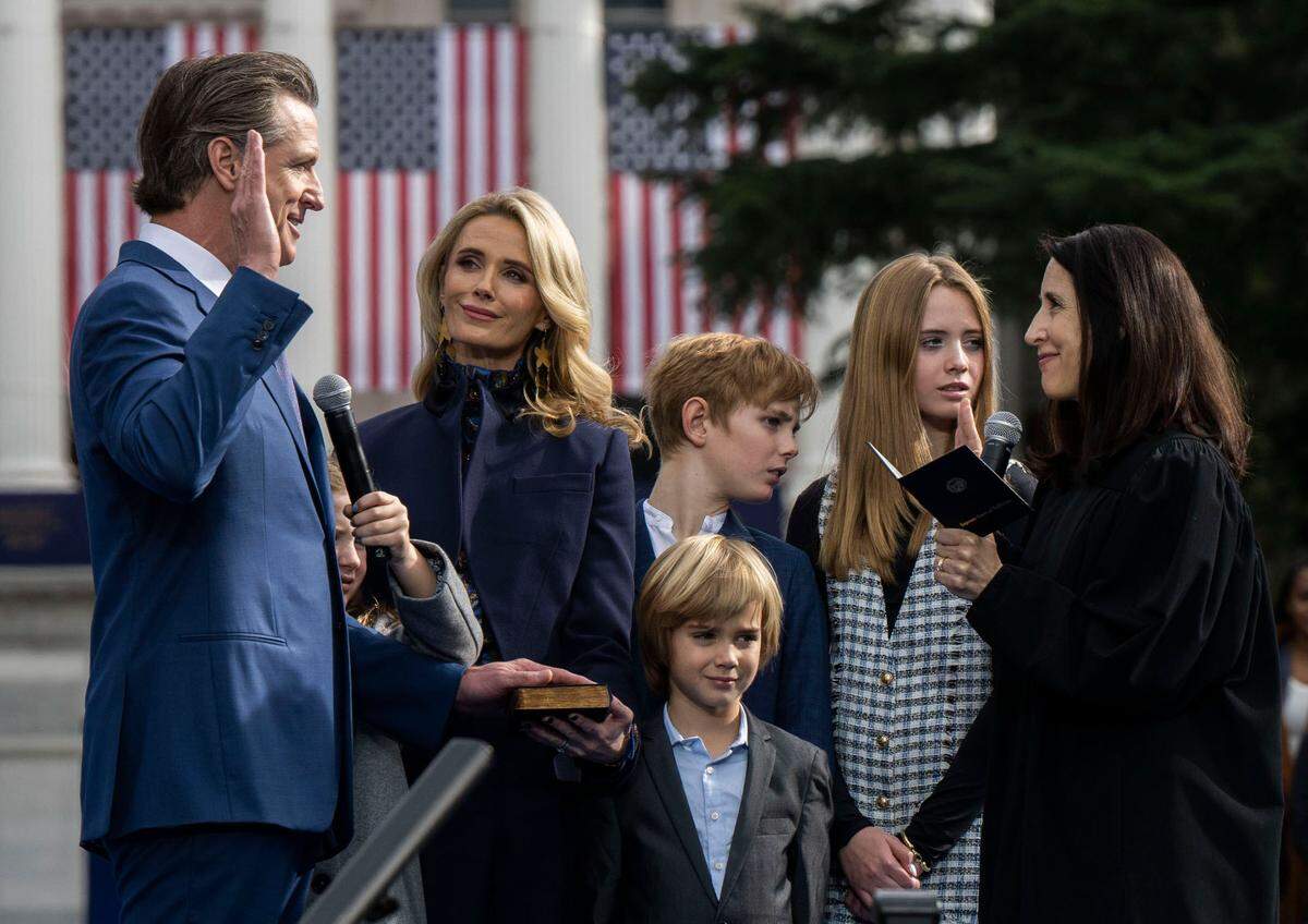 Gov. Gavin Newsom is sworn in during his inauguration Friday by new Chief Justice Patricia Guerrero. His children look at a heckler who held a sign opposing COVID-19 vaccines and supporting Ron DeSantis for President in 2024.
