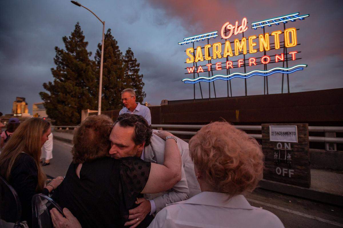 Terry Harvego, center, embraces his mother, Sandy Harvego, on the I Street viaduct after a ceremonial first lighting of a neon-alternative LED Old Sacramento Waterfront sign Wednesday, June 22, 2022, seen rising more than 20 feet above the California State Railroad Museum rooftop. Terry’s father, Lloyd Harvego, who died June 13, was a longtime proponent of the sign, designed by Sacramento-based Pacific Neon Company and funded by the Downtown Sacramento Foundation. “To say were grateful for Lloyd and his vision is an understatement,” said Michael Ault, standing behind them, executive director of the Downtown Sacramento Partnership. “I will miss him personally, and thank his family very much for the work that they did. “