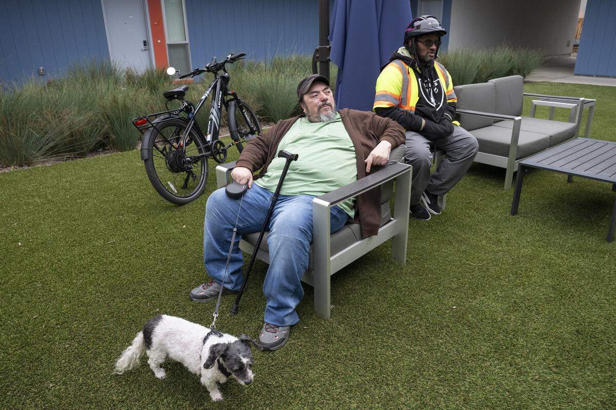 AJ Ortiz, 61, left, and Dwayne McDaniel, 53, sit in the courtyard of their apartment building in Sacramento on Thursday, as Ortiz’s dog, Lacy, stands near them. Ortiz and McDaniel were both beneficiaries of an e-bike voucher program.