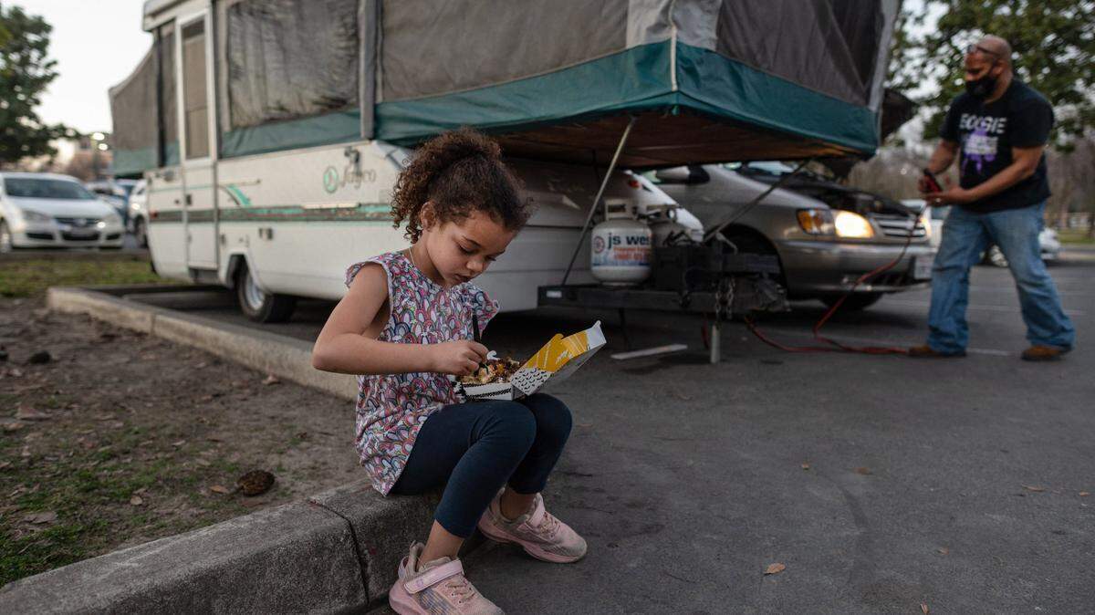 Shyann Slain, 7, eats a meal from Del Tacos as her father Anthony Slain, 45, uses his van’s battery to generate electricity as he set up a camper for them to sleep that night in the South Natomas Community Park parking lot on Saturday, Jan.17, 2021. The parents say they usually leave the car running all night and sleep in shifts to keep the children warm.