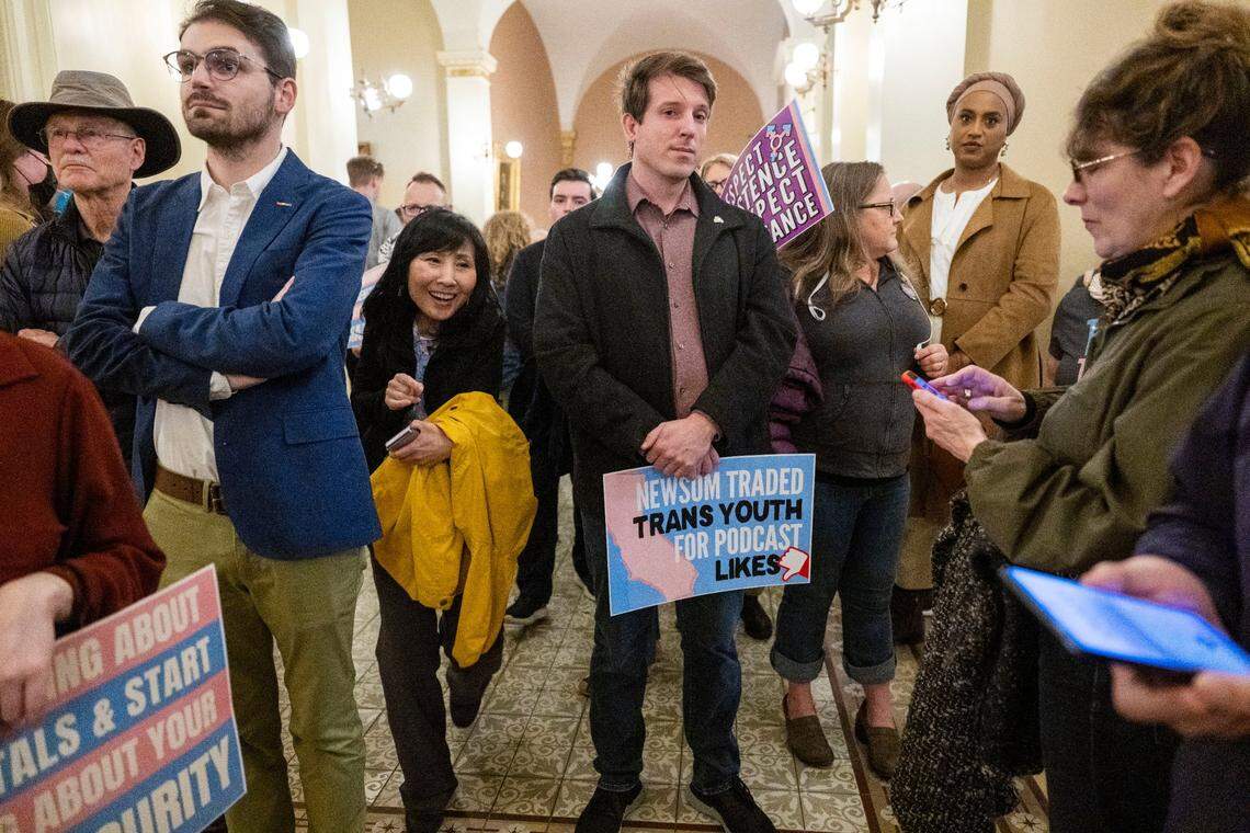 A man holds a sign that says “Newsom traded trans youth for podcast likes” as people wait to make make public comments on Assembly Bill 844 by Bill Essayli, R-Corona, during an Assembly committee hearing on April 1 at the state Capitol. The bill would would reverse a 2013 law that allows California students to use locker rooms and play on sports teams that reflect their gender identity.