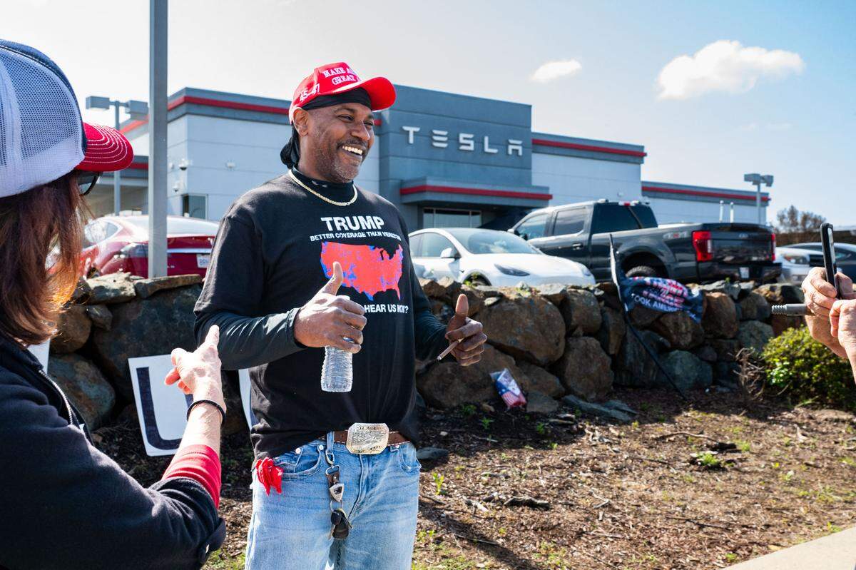 Klifton Knowling, who likes to go by the nickname “Black MAGA,” smiles as a fellow counter protester takes a photo of his “Trump: Better coverage than Verizon” T-shirt in front of the Tesla dealership as anti-Tesla protesters demonstrate across the street Saturday, March 29, 2025.