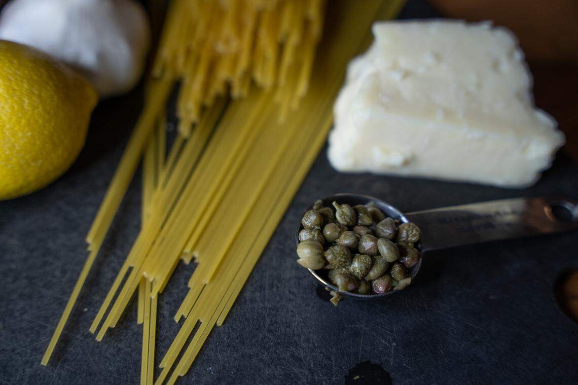 Capers in a measuring spoon, alongside butter and dried pasta.
