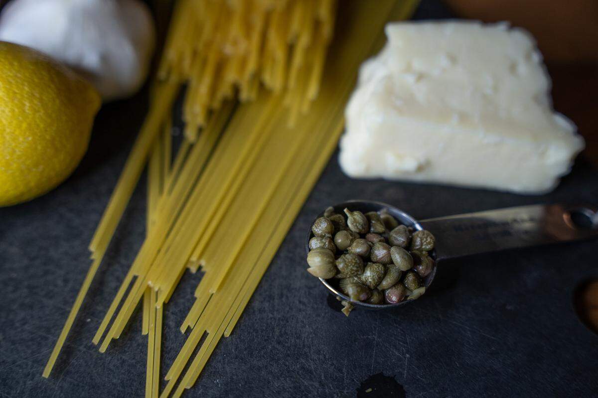 Capers in a measuring spoon, alongside butter and dried pasta.